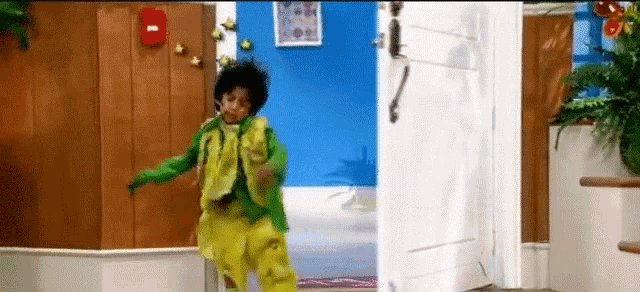 a young boy in a green jacket and yellow pants is dancing in an open doorway .