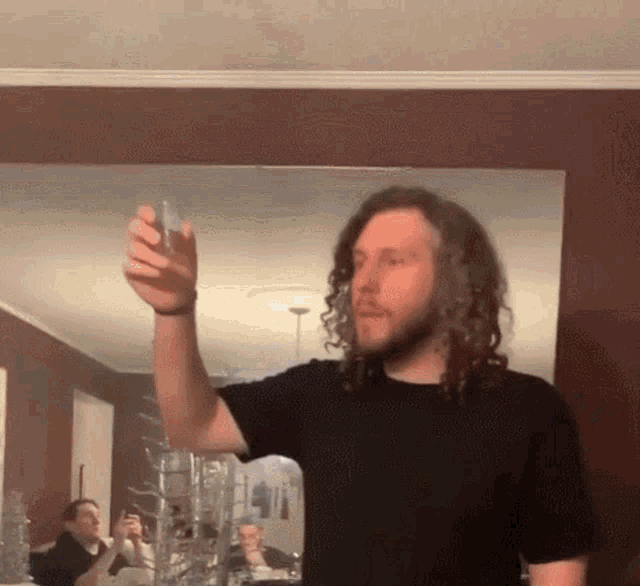 a man with long curly hair is holding a glass in front of a mirror in a living room .
