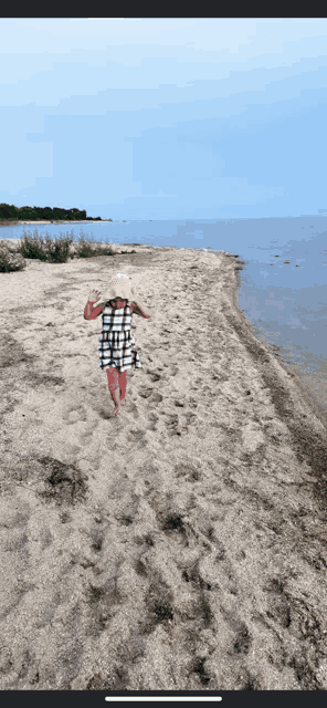 a little girl wearing a plaid dress and a hat is walking on a sandy beach