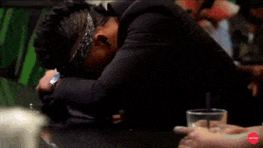 a woman sits at a table with a drink and a man laying on the table