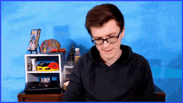 a young man wearing glasses sits in front of a blue background