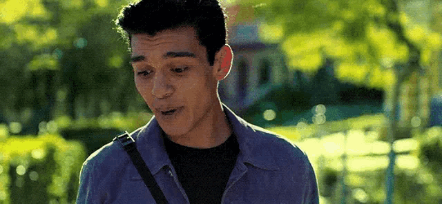 a man in a blue shirt is standing in a park with a shoulder bag .