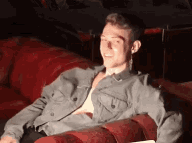 a man is sitting on a red couch with his shirt off and smiling .