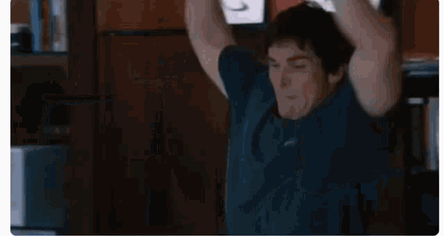 a man in a blue shirt is standing in a room with his hands behind his head .