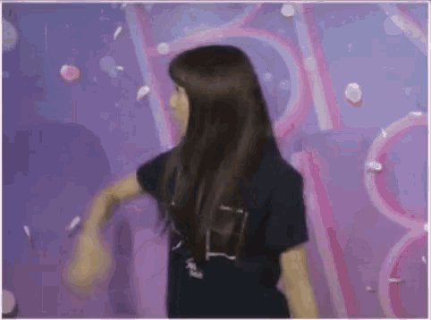 a woman with long hair is dancing in front of a purple wall with flowers falling from the ceiling .