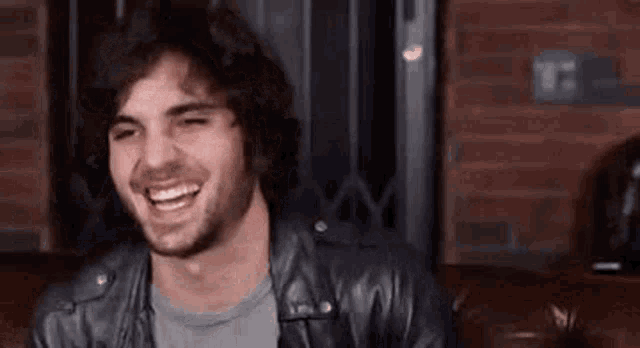 a man with a beard and a leather jacket is sitting on a couch and smiling .
