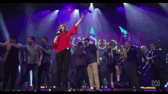 a woman in a red shirt sings into a microphone in front of a sign that says festival