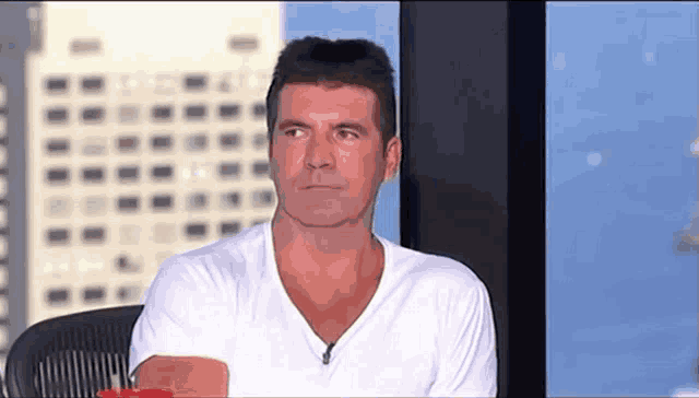 a man in a white t-shirt is sitting in front of a window with a building in the background .