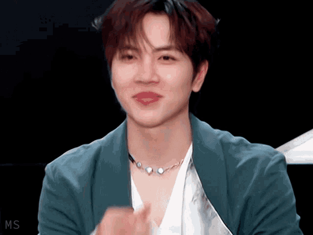 a young man wearing a necklace and a green jacket is clapping