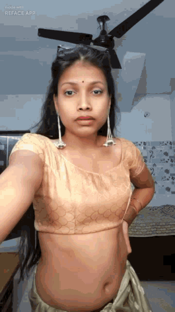a woman in a crop top and earrings is taking a selfie in a room with a ceiling fan .
