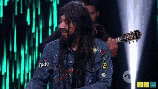 a man with long hair and a beard is wearing a denim jacket and holding a guitar on a stage .