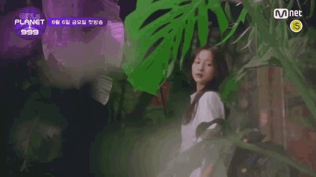 a woman in a white dress is standing in front of a plant with a mnet logo behind her .