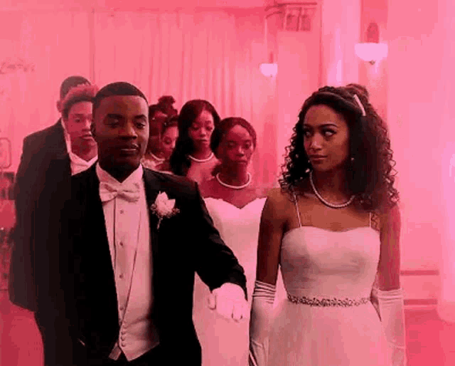 a man in a tuxedo and a woman in a white dress are walking down a red carpet with other people .
