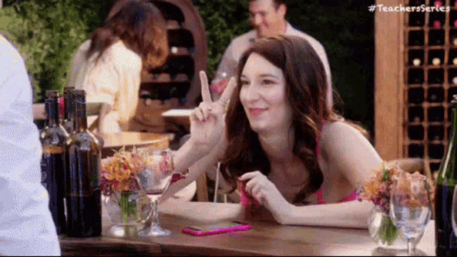a woman is sitting at a table with wine glasses and bottles of wine and giving a peace sign .