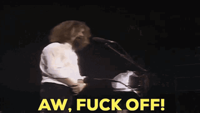 a man with long hair and a beard is standing in front of a microphone and says aw fuck off