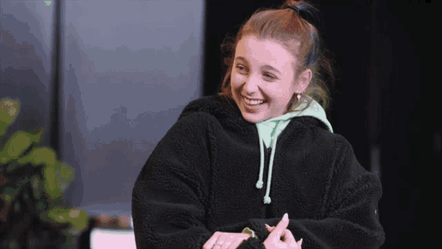 a young woman wearing a black jacket and a green hoodie smiles