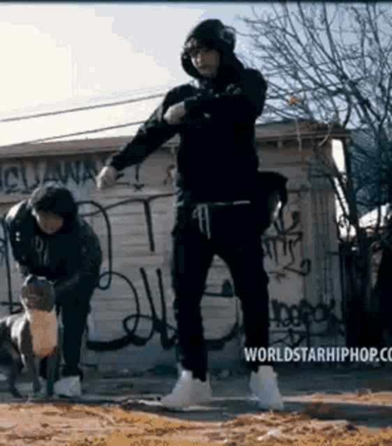 a man in a black hoodie is standing next to a dog in front of a building with graffiti on it .