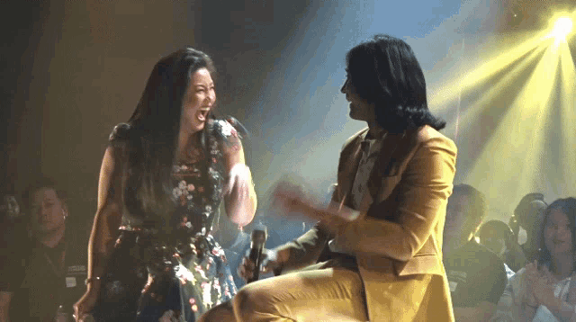 a woman in a floral dress laughs while a man in a yellow suit sits on a stage