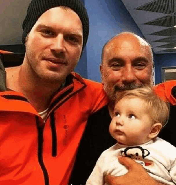 two men are holding a baby in their arms . one of the men is wearing an orange jacket .