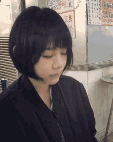 a girl with short black hair is sitting in front of a wall with asian writing on it