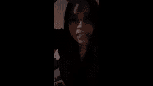 a woman with long black hair is sitting in a dark room talking on a cell phone .
