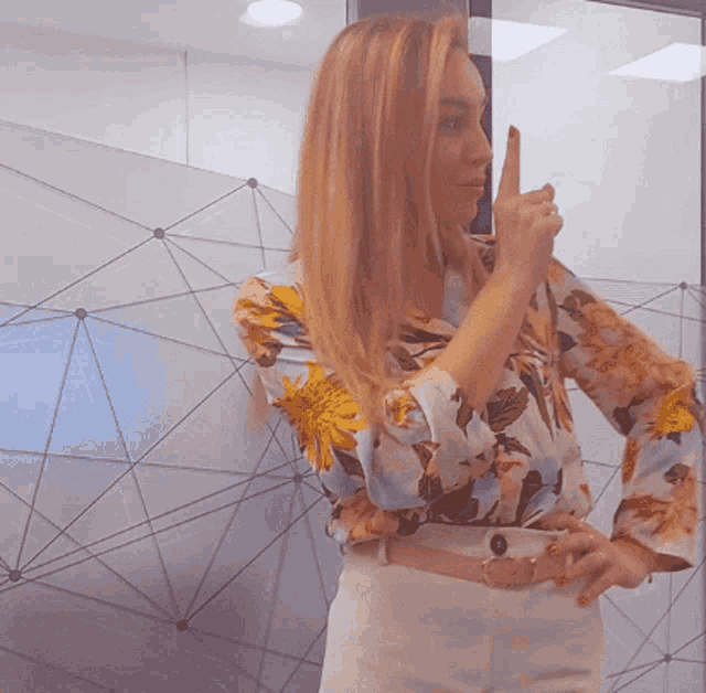 a woman wearing a floral shirt and white pants holds her finger to her lips