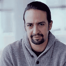 a man with a beard and long hair is wearing a gray sweater and looking at the camera .