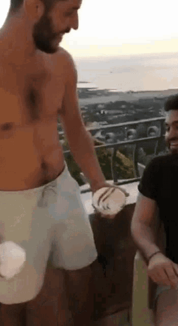 a man without a shirt is holding a bowl of food while another man sits next to him .
