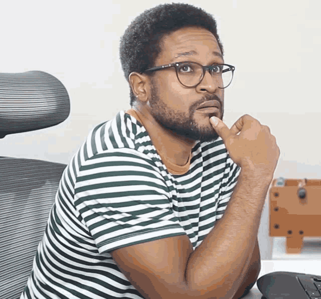 a man wearing glasses and a striped shirt is sitting at a desk with his hand on his chin
