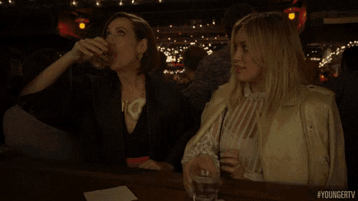 two women are sitting at a bar holding shot glasses and pointing at something .