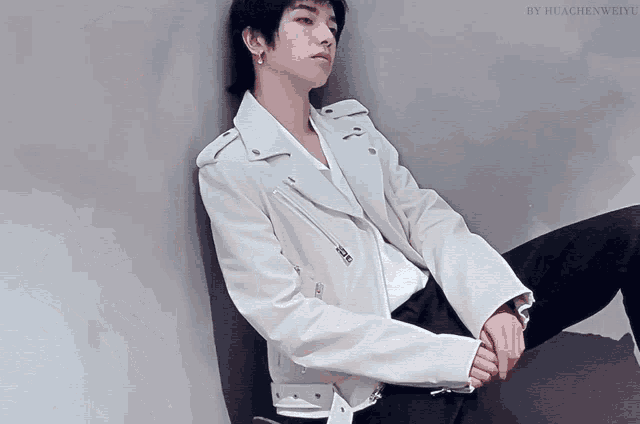 a young man leaning against a wall wearing a white jacket and black pants