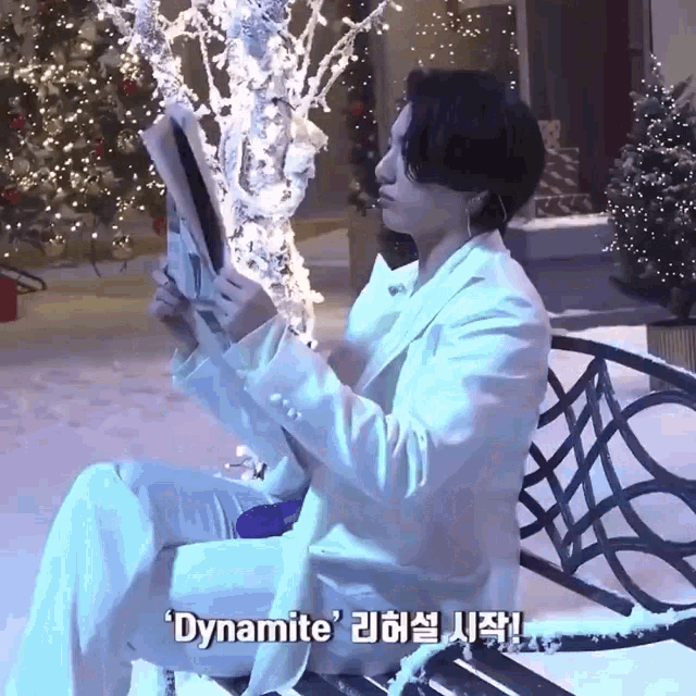 a man in a white suit is sitting on a bench reading a newspaper with dynamite written on the bottom