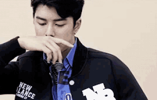 a young man wearing a new balance jacket and tie is covering his nose with his hand .