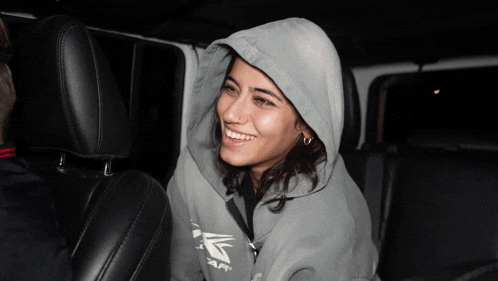 a woman wearing a grey adidas hoodie smiles while sitting in the back seat of a car