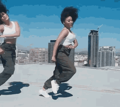 two women are dancing on a rooftop in front of tall buildings