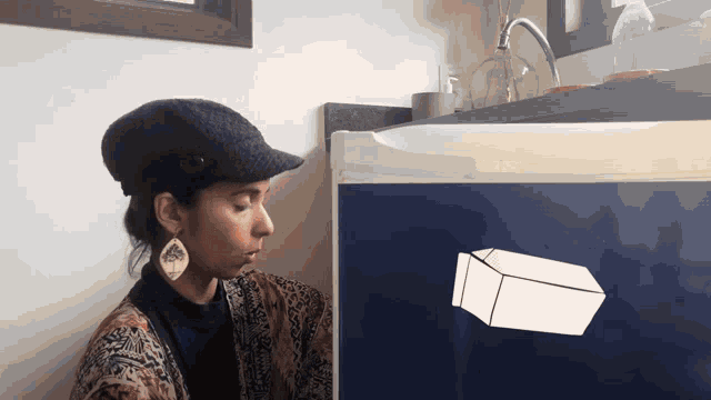 a woman wearing a hat and earrings sits in front of a blue refrigerator