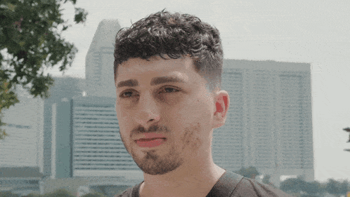 a man with curly hair and a beard looks at the camera with a city skyline in the background