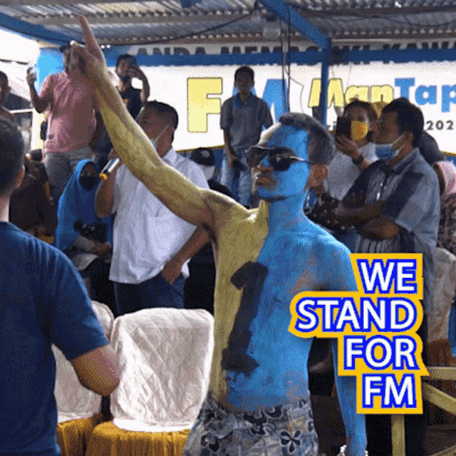 a man with blue paint on his body is standing in front of a crowd and a sign that says we stand for fm