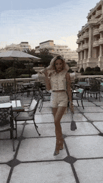 a woman in a crop top and shorts stands in front of a patio with tables and chairs