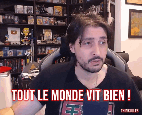 a man with a beard says " tout le monde vit bien " in front of a bookshelf