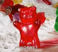 a red gummy bear is standing in front of a pile of candy and says `` not yet '' .