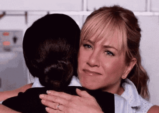 a woman is hugging another woman in a kitchen . the woman is wearing a ring on her finger .