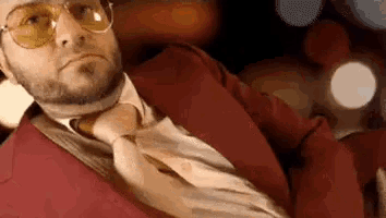 a man in a red suit and tie is sitting in a car with his legs crossed .