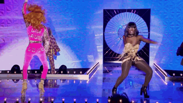 two drag queens are dancing on a stage with a fan in the background .