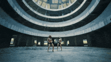two women are dancing in an empty building with a dome in the background