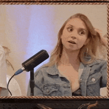 a woman in a denim jacket is standing in front of a microphone and looking at the camera .