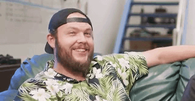 a man with a beard is sitting on a bean bag chair wearing a hat and a hawaiian shirt .