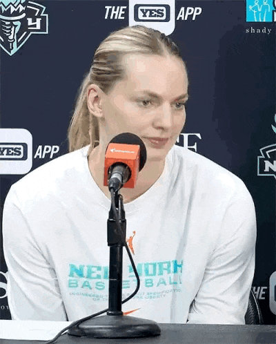 a woman wearing a white shirt that says new york basketball is sitting in front of a microphone .