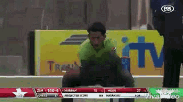 a man in a green shirt is squatting down on a field in front of a fox tv advertisement .