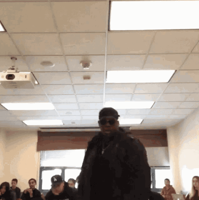a man wearing sunglasses and a ny hat stands in front of a group of people in a classroom